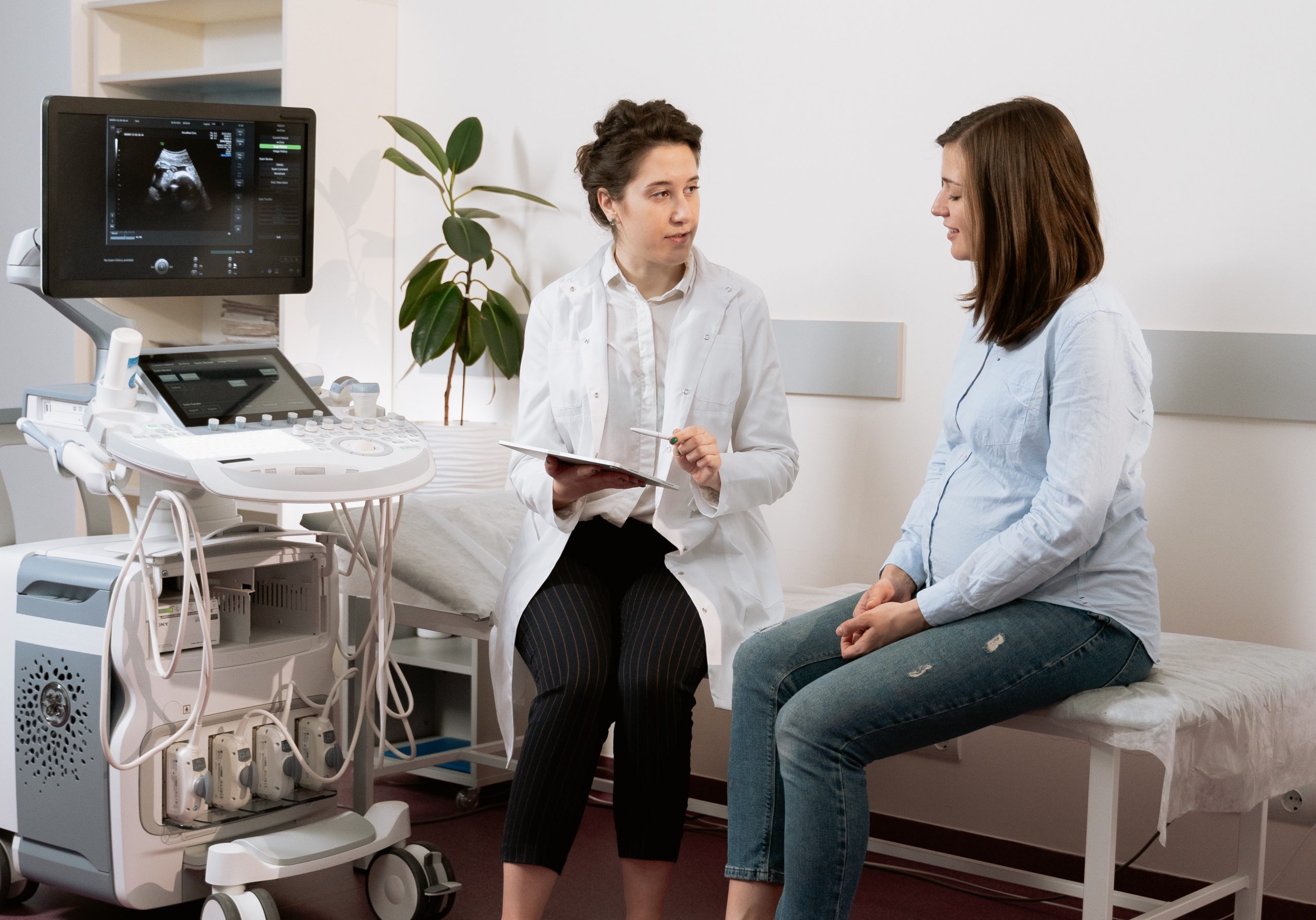 Eine Ärztin sitzt neben einer Patientin auf einer Untersuchungsliege. Sie hält ein Tablett und einen Stift und erklärt ihrer schwangeren Patientin etwas. Links neben den beiden steht ein Ultraschallgerät.